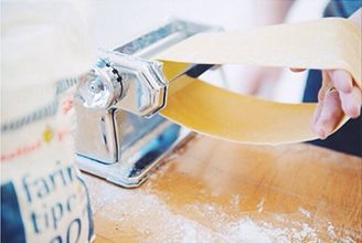 Rolling a sheet of fresh pasta through a hand-cranked machine beside a bag of tipo 00 flour