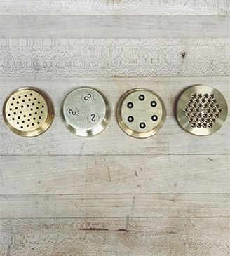 Four brass pasta dies, each cutting a different shape, laid on a wooden board