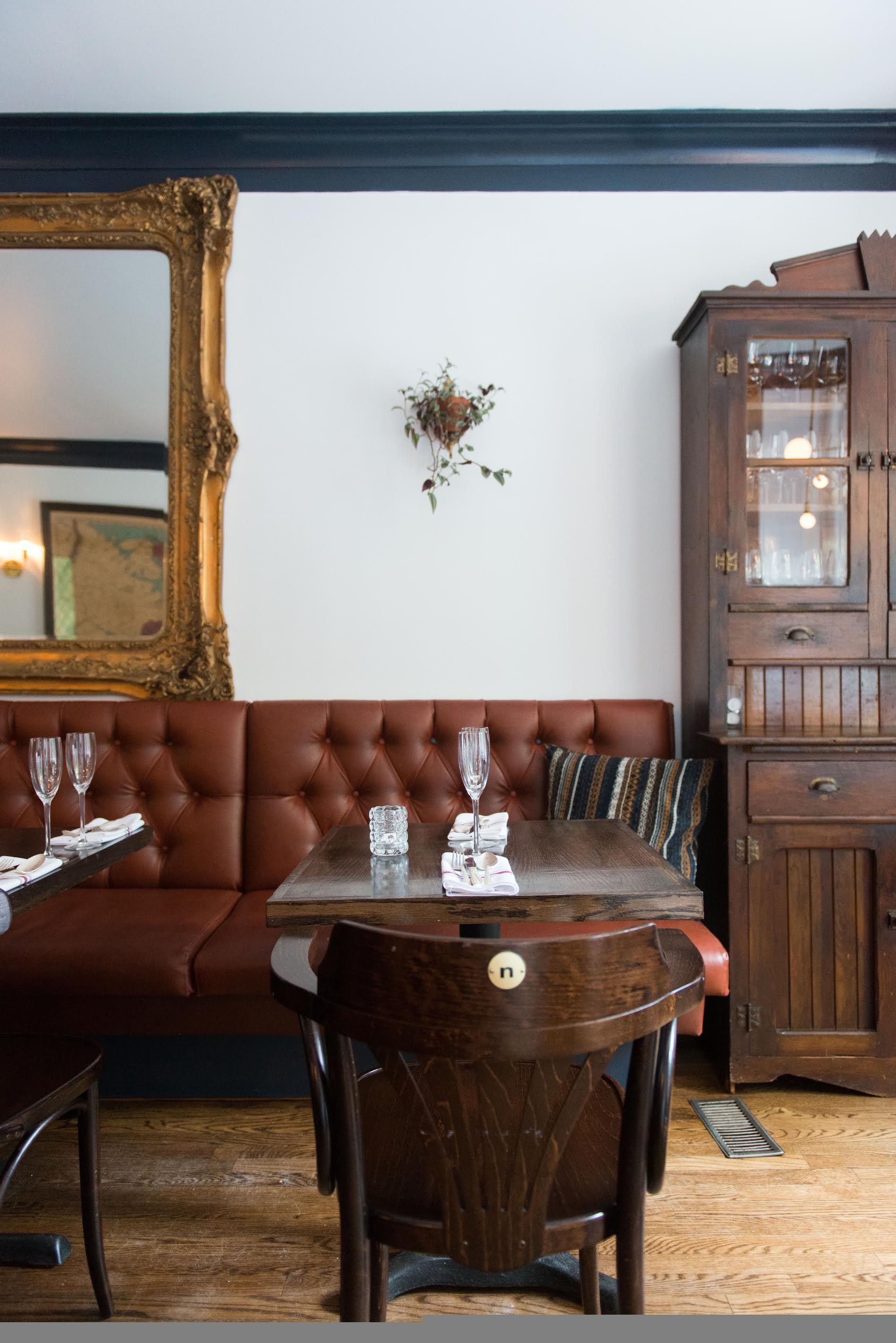 Dining room banquette in oxblood leather beneath a gilt mirror, with an antique glass-front hutch and a table set for two
