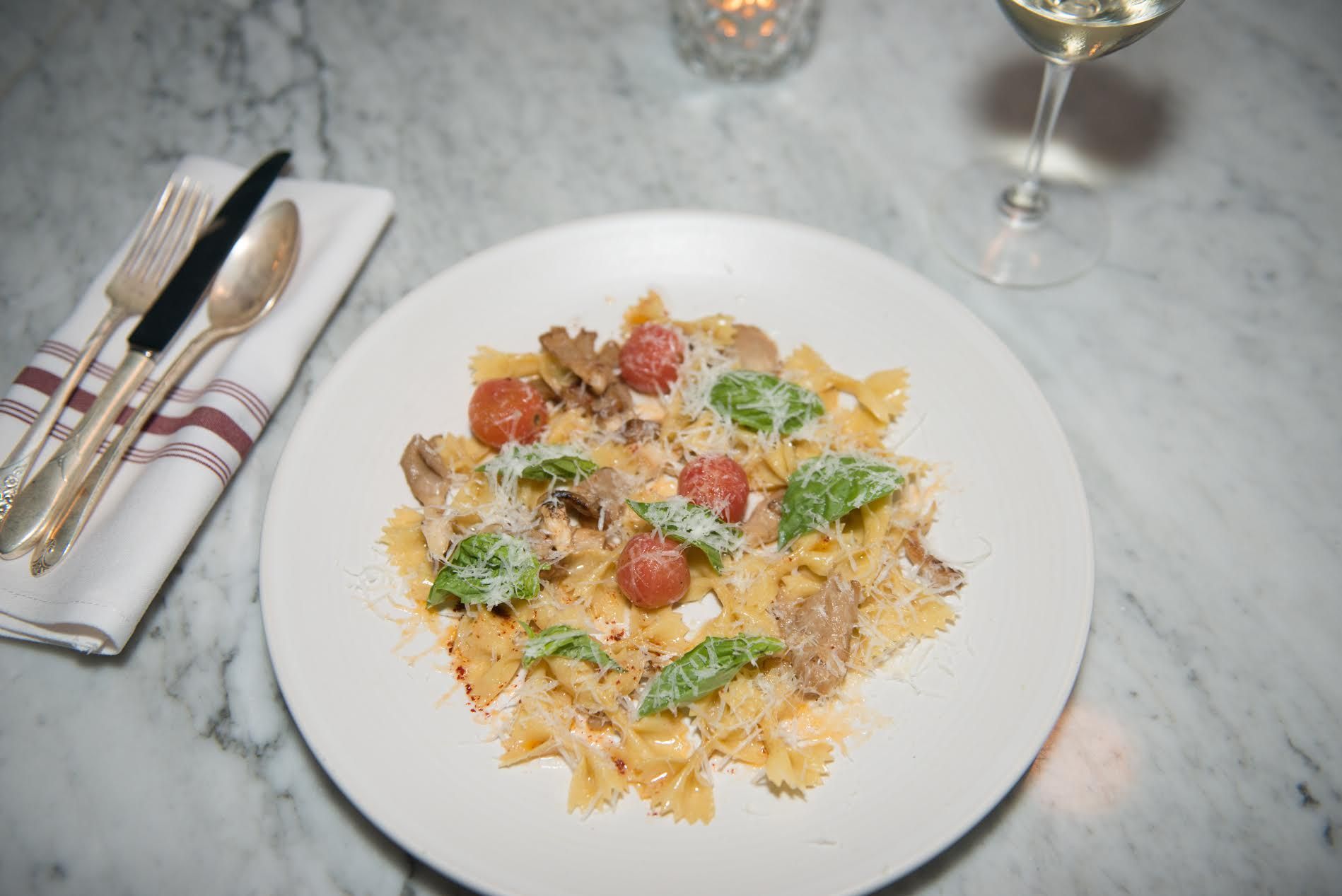 Farfalle with cherry tomatoes, mushrooms, basil and shaved parmesan on a marble table beside a glass of white wine