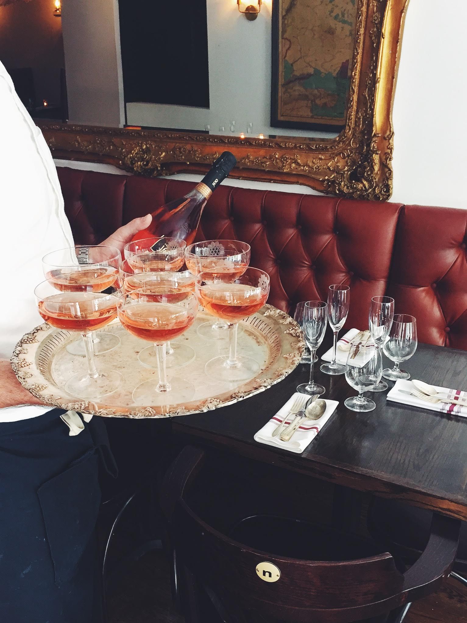 A server carrying a silver tray of coupes filled with sparkling rose past the dining room banquette